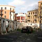Al centro spicca lo scheletro di un�auto bruciata al cui fianco si trova un cane randagio. Alle spalle gli alti e colorati palazzi del quartiere Madonnella 